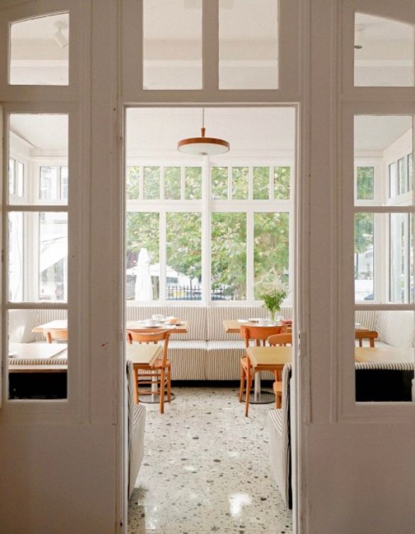 View through French doors into a bright sunlit room with wooden chairs and terrazzo flooring.