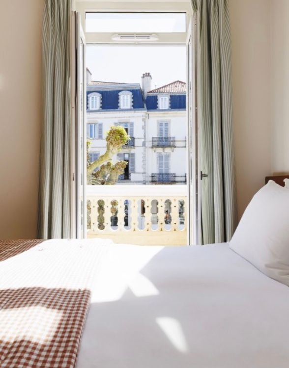 A bright bedroom with white bedding, gingham pillows, and large windows overlooking a scenic balcony view.