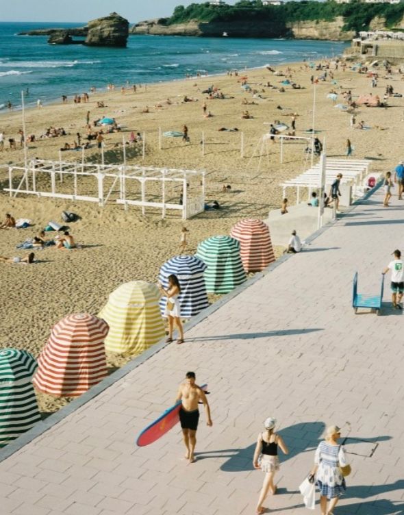 Colorful striped beach umbrellas line a sandy beach walkway with beachgoers enjoying the sunny summer day.
