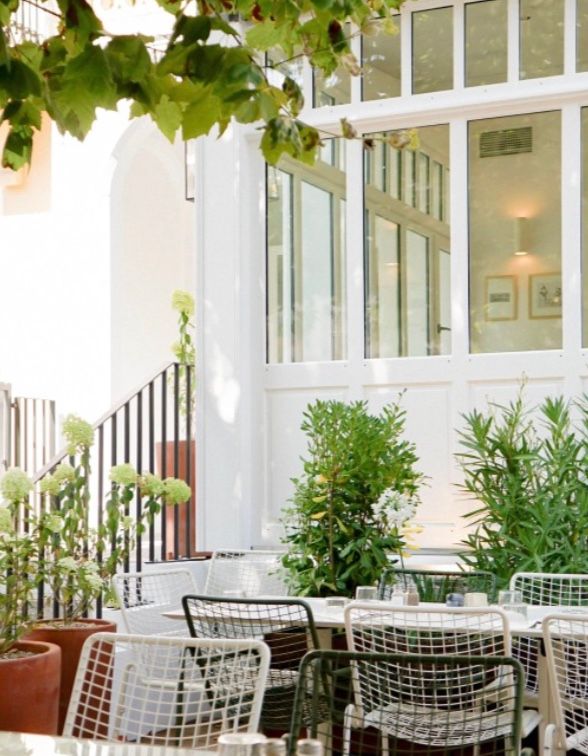 Outdoor restaurant terrace with white wire chairs and lush greenery framing a sunlit glass-enclosed dining space.