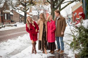 Four people smiling and laughing together on a snowy street decorated for Christmas, wearing colorful winter outfits.