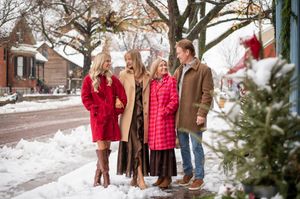 Family of four smiling outdoors in winter coats on a snowy small-town street decorated for Christmas.