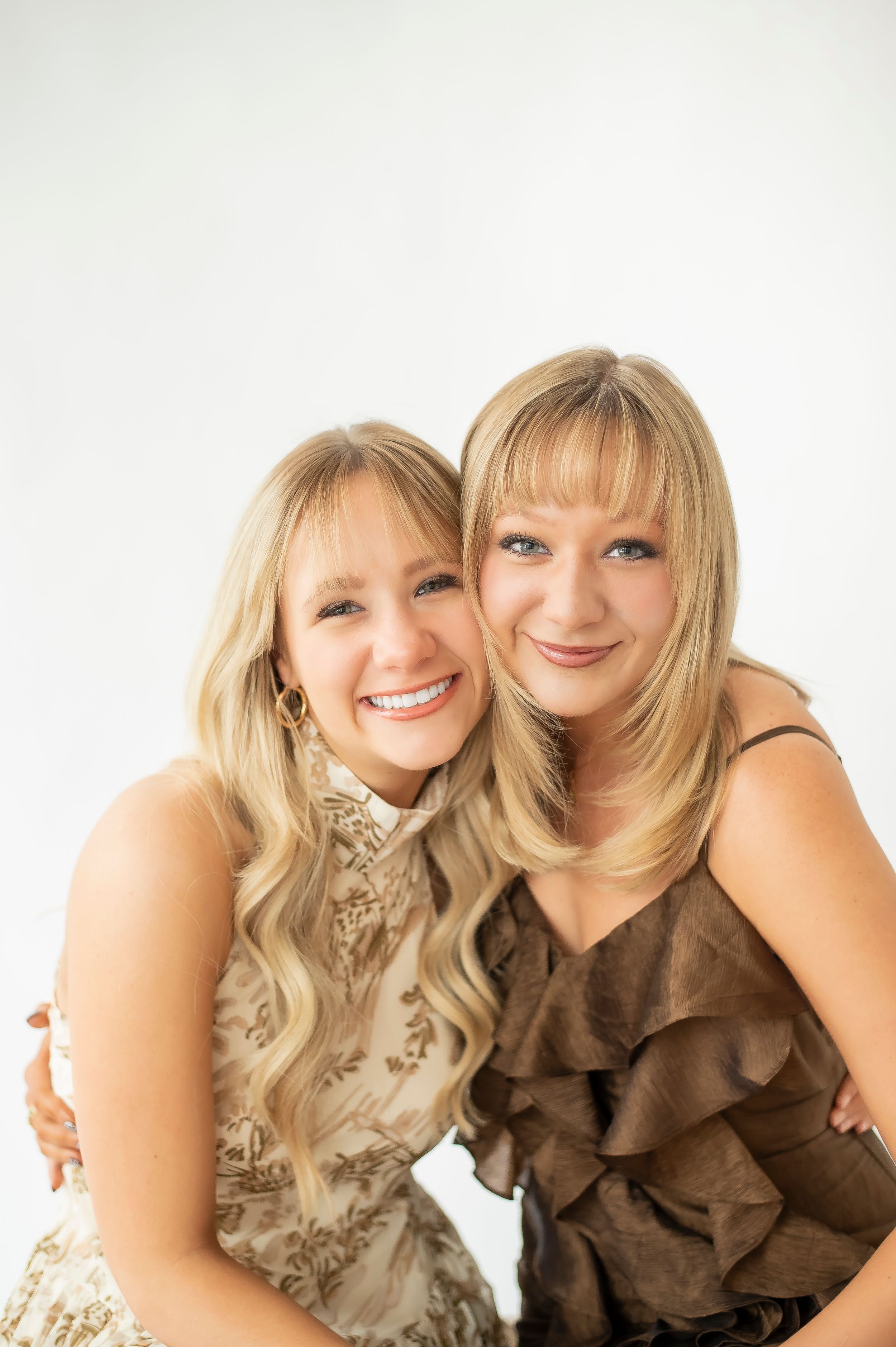 Two smiling blonde women embracing closely together in a bright white photography studio setting.