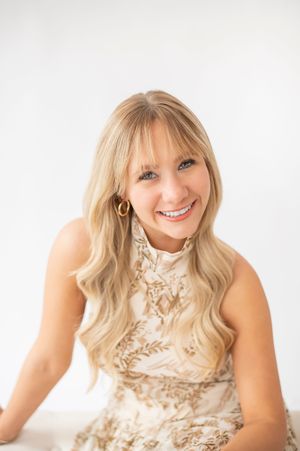 Smiling blonde woman with wavy hair wearing a gold and white floral dress in a bright white studio.