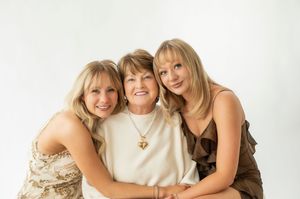 Three blonde women smile warmly together in a studio portrait, wearing elegant earth-tone outfits.