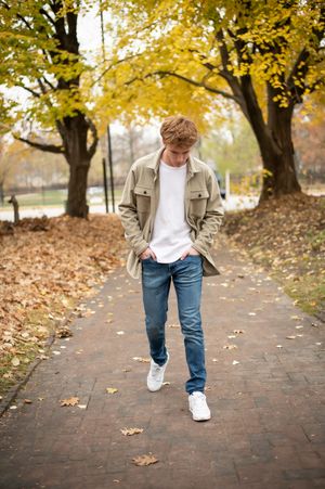 Young man in beige jacket and jeans walking on brick path surrounded by fall foliage.