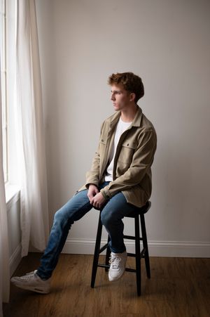 Young man in a tan jacket and jeans sits on a stool gazing out a window in natural light.