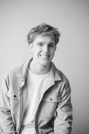 Smiling young man with curly hair wearing a beige jacket poses for a black and white portrait.