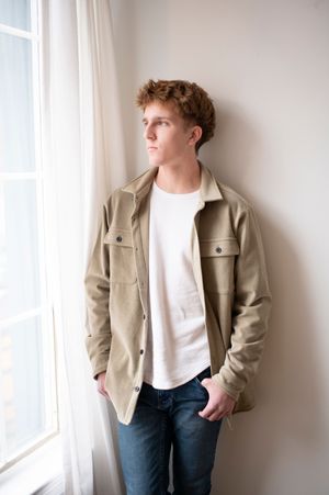 Young man in a beige jacket and jeans leans against a window frame in soft natural light.