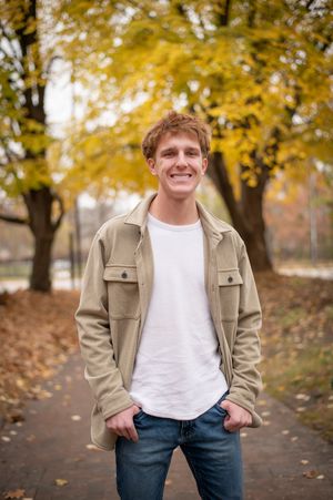 Smiling young man in beige jacket and white tee standing on autumn park path with yellow trees.