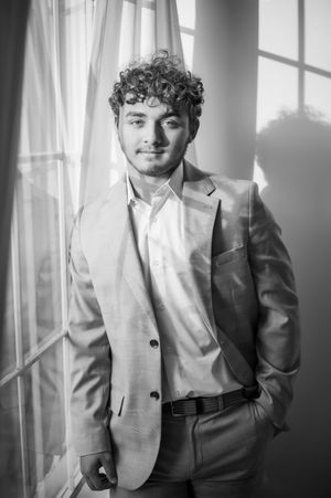 Black and white portrait of a curly-haired young man in a suit standing by a window.
