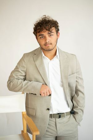 Confident young man in beige blazer leaning on white chair against neutral background.