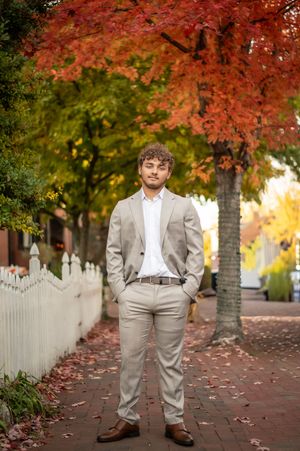 Young man in beige suit standing on brick sidewalk surrounded by vibrant red and orange autumn trees.