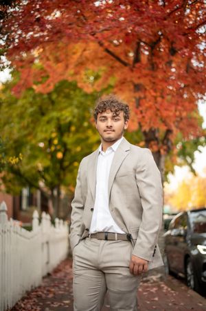 Young man in beige suit poses on autumn sidewalk with vibrant red and orange foliage trees in background.