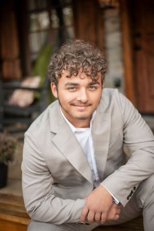 Young man with curly hair wearing a beige suit smiles confidently outdoors near a rustic wooden building.