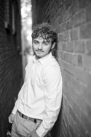 Young man with curly hair leaning against brick wall in alley, wearing white shirt, black and white photo.