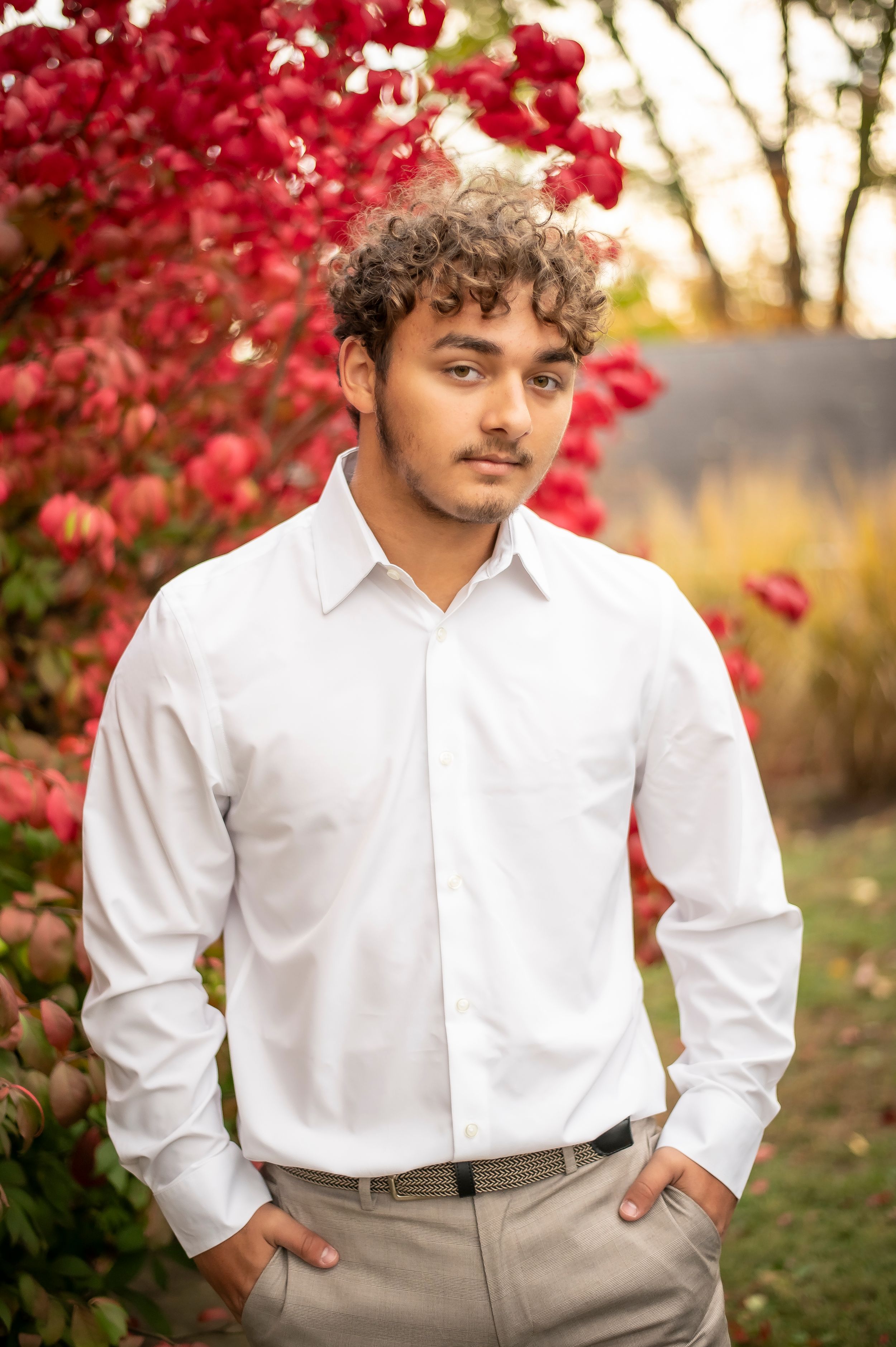 Young man with curly hair in white dress shirt stands before vibrant red autumn foliage in outdoor portrait.