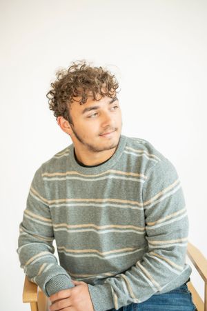 Young man with curly hair in striped green sweater sitting casually against white background.