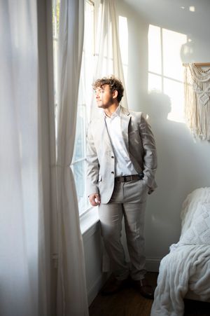 Stylish young man in gray suit standing by sunlit window in bright bedroom interior.