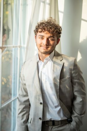 Smiling young man with curly hair in gray blazer posing by a sunlit curtained window.