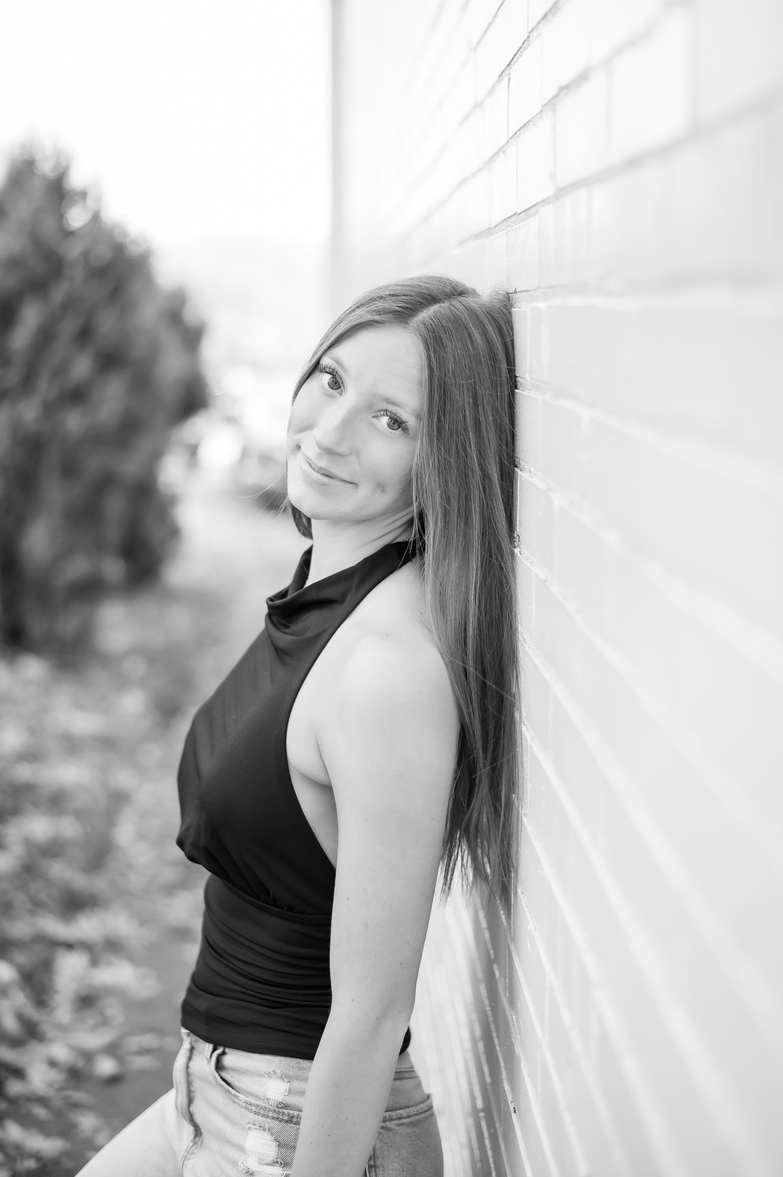 Black and white portrait of a smiling young woman leaning against a brick wall outdoors.