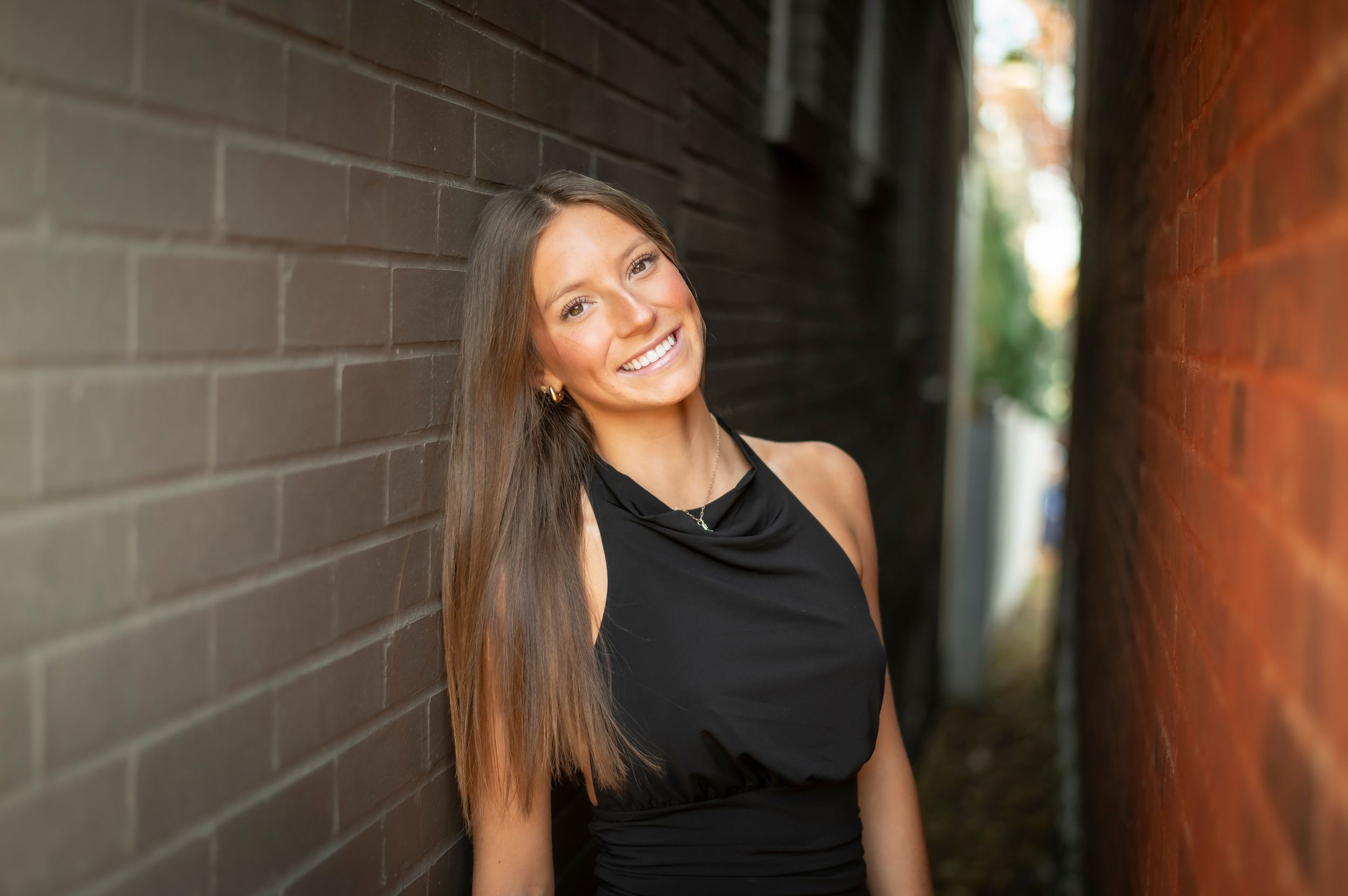 Young woman with long brown hair smiling in a black sleeveless dress leaning against a brick wall.