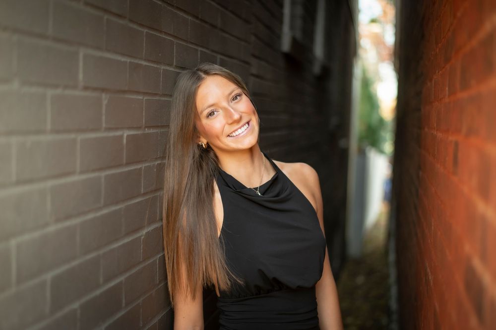 Young woman with long brown hair smiling in a black sleeveless dress leaning against a brick wall.