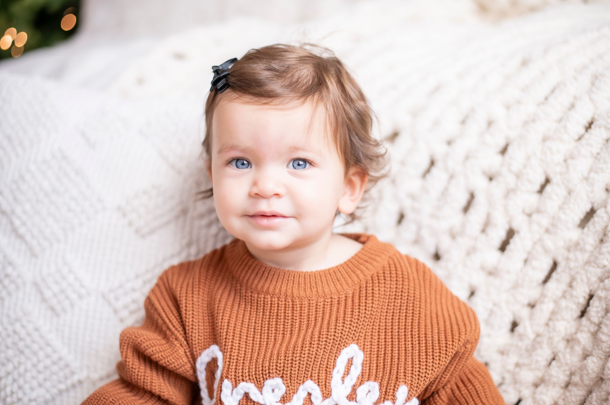 Adorable baby with blue eyes wearing an orange knit sweater sitting against a white textured background.