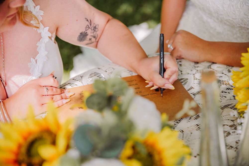 LGBTQ+ Dorset Wedding at the Gaggle of Geese, Tipi wedding venue in Dorset. Bride signs the marriage certificate surrounded by sunflowers