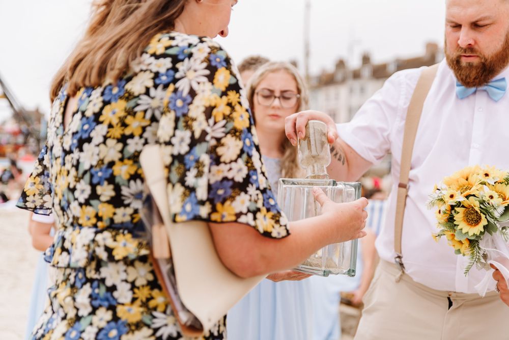 UK beach wedding on Weymouth Beach with Ceremonies by Laura Bell. Celebrant helps bride with dress. Sunflower wedding bouquet
