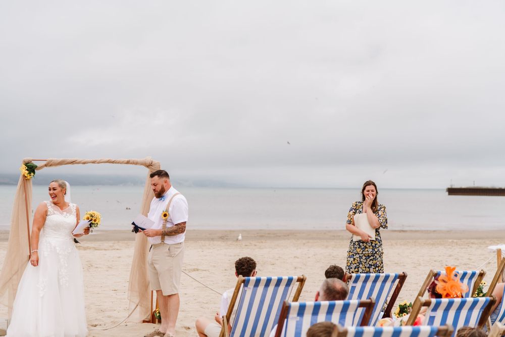 UK beach wedding on Weymouth Beach with Ceremonies by Laura Bell. Celebrant helps bride with dress. Sunflower wedding bouquet