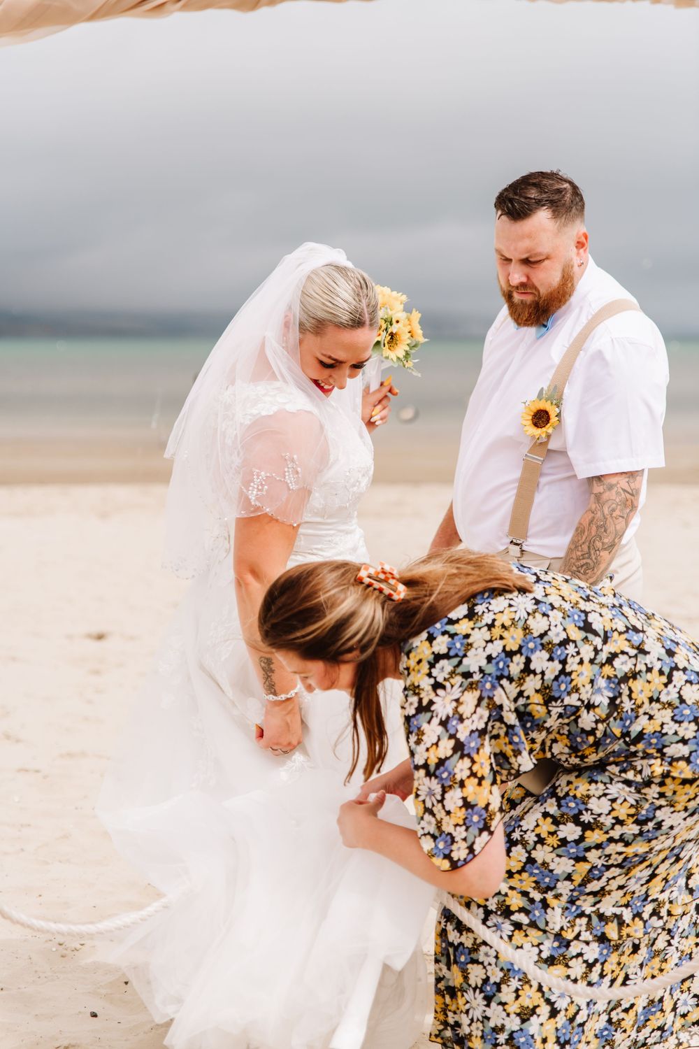 UK beach wedding on Weymouth Beach with Ceremonies by Laura Bell. Celebrant helps bride with dress. Sunflower wedding bouquet