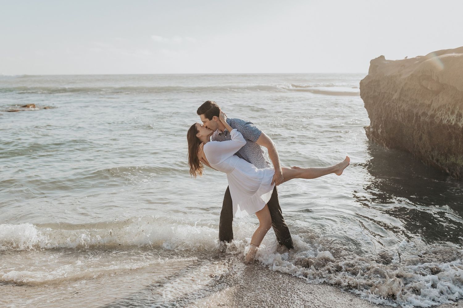 Couple embracing on rocky beach during sunset as ocean waves crash around them.