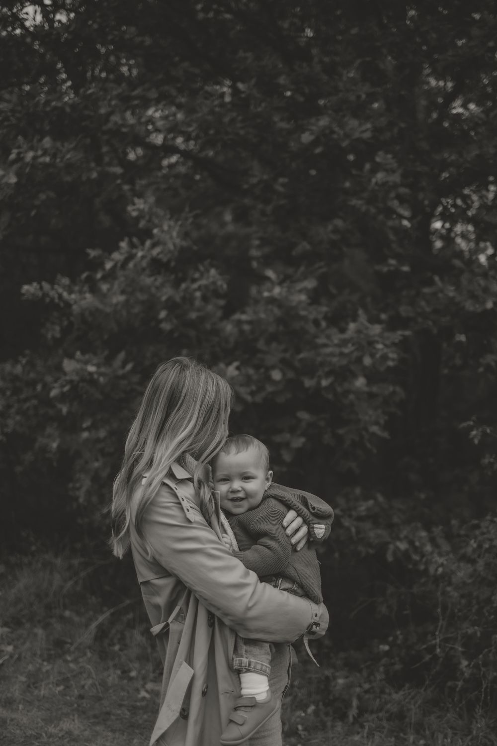 Monochrome image of a Mum embracing a smiling baby outdoors against foliage.