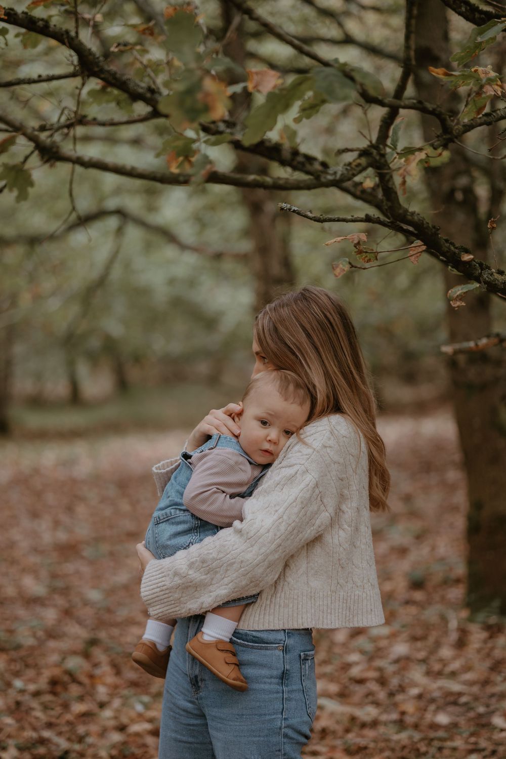 Mum in cable knit sweater holding sleeping baby under fall trees with scattered leaves.