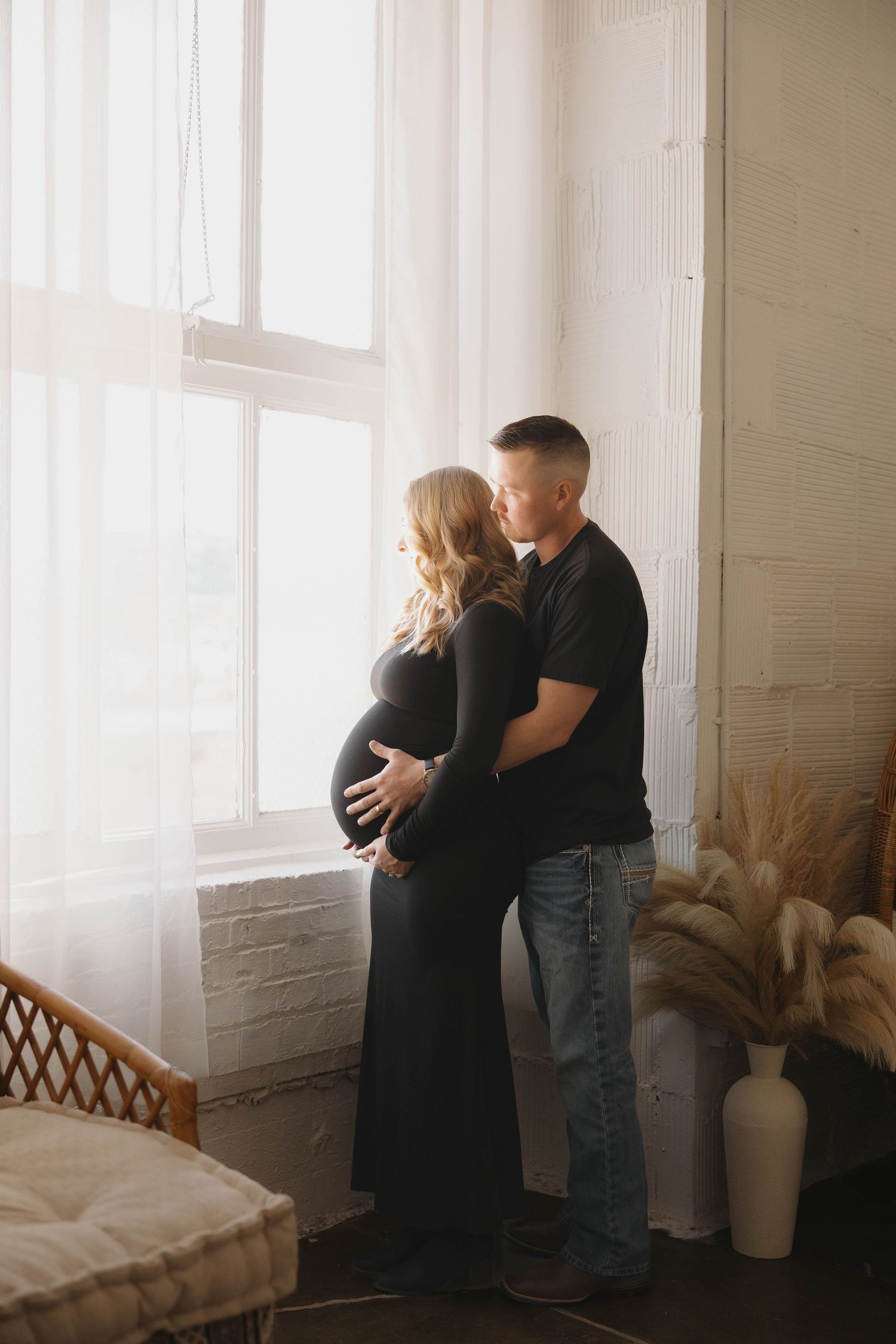A couple embraces in a bright white room with sunlit windows and minimal decor.