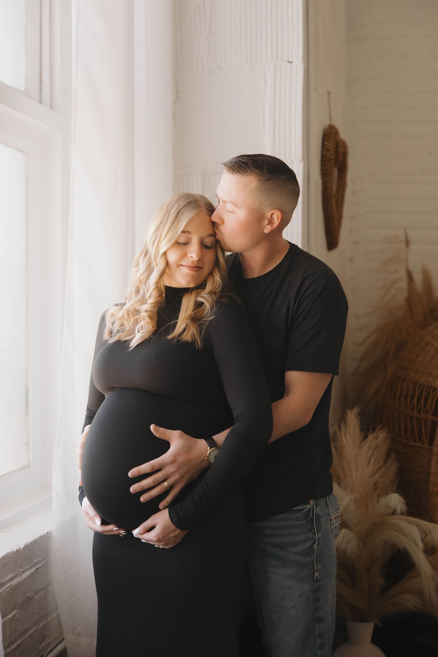 Intimate maternity photo series showing couple embracing in front of bright window with pampas grass decor.