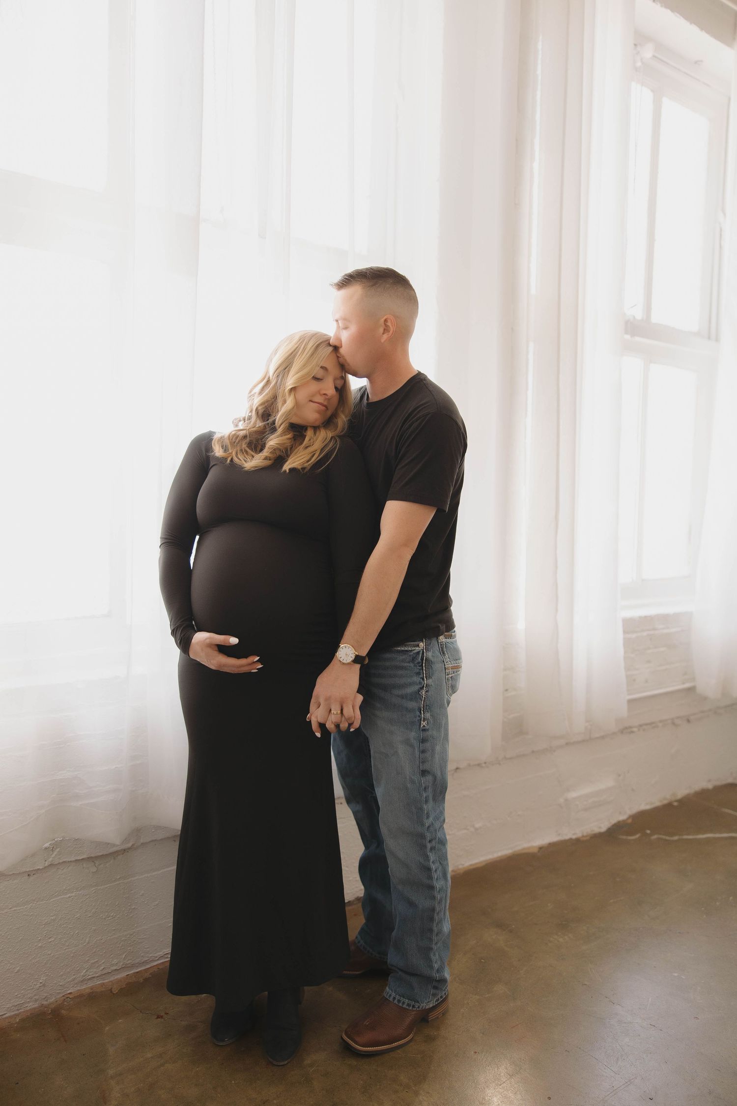 Couple shares intimate moment in bright room as woman in black dress shows off baby bump during maternity photoshoot.