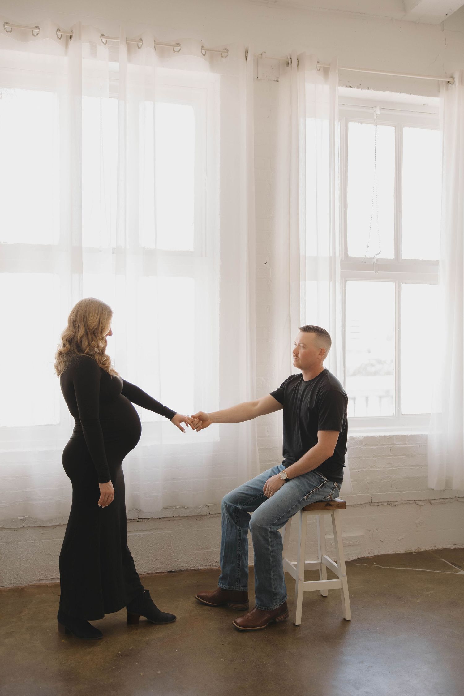 Couple holds hands in intimate maternity photo with sheer white curtains and natural light in minimal studio setting.