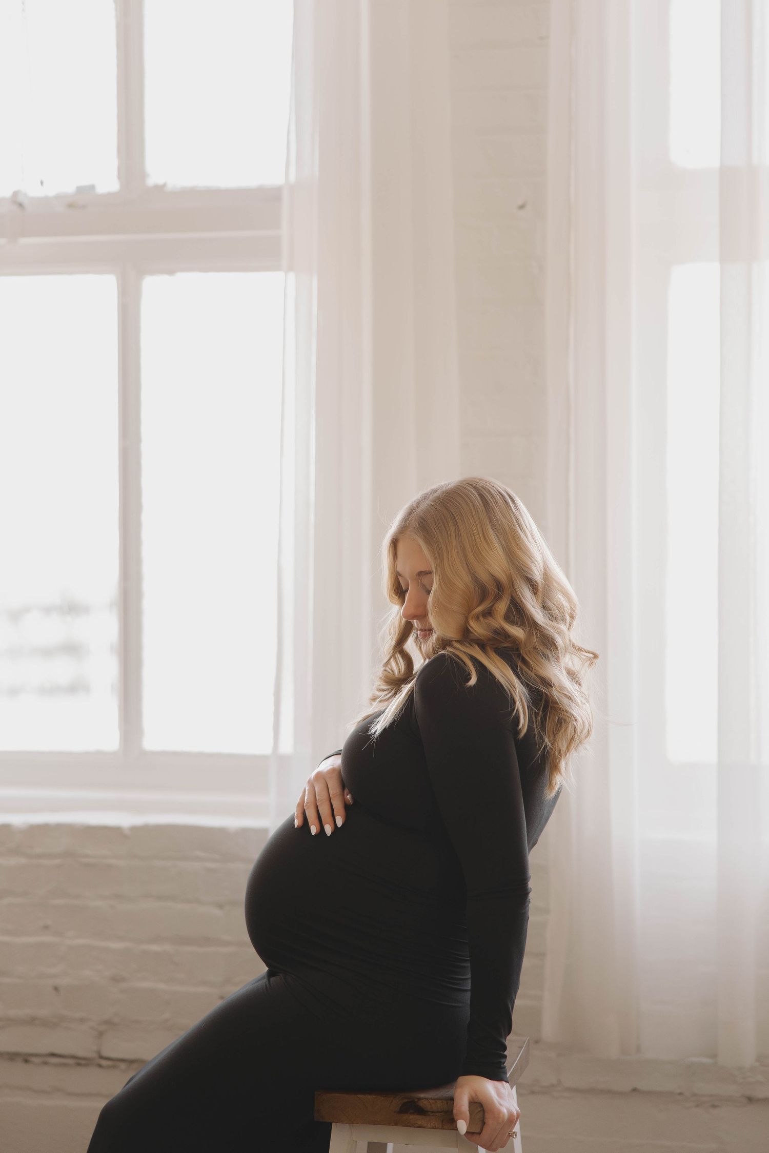 Woman in black sweater sits alone by bright window in minimalist white room showing different contemplative poses.