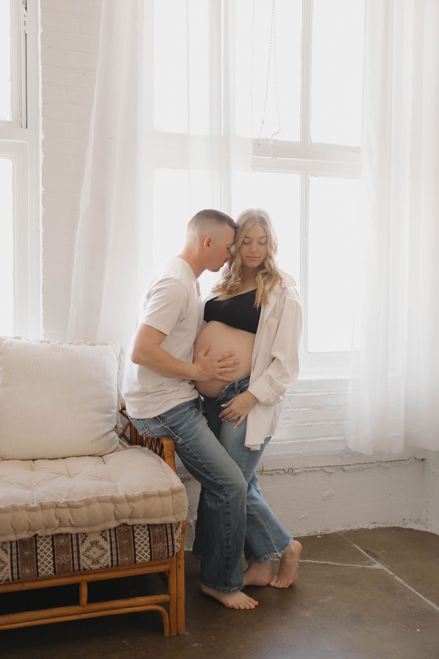 A couple shares an intimate moment on a cozy daybed with white curtains and natural light streaming through windows.