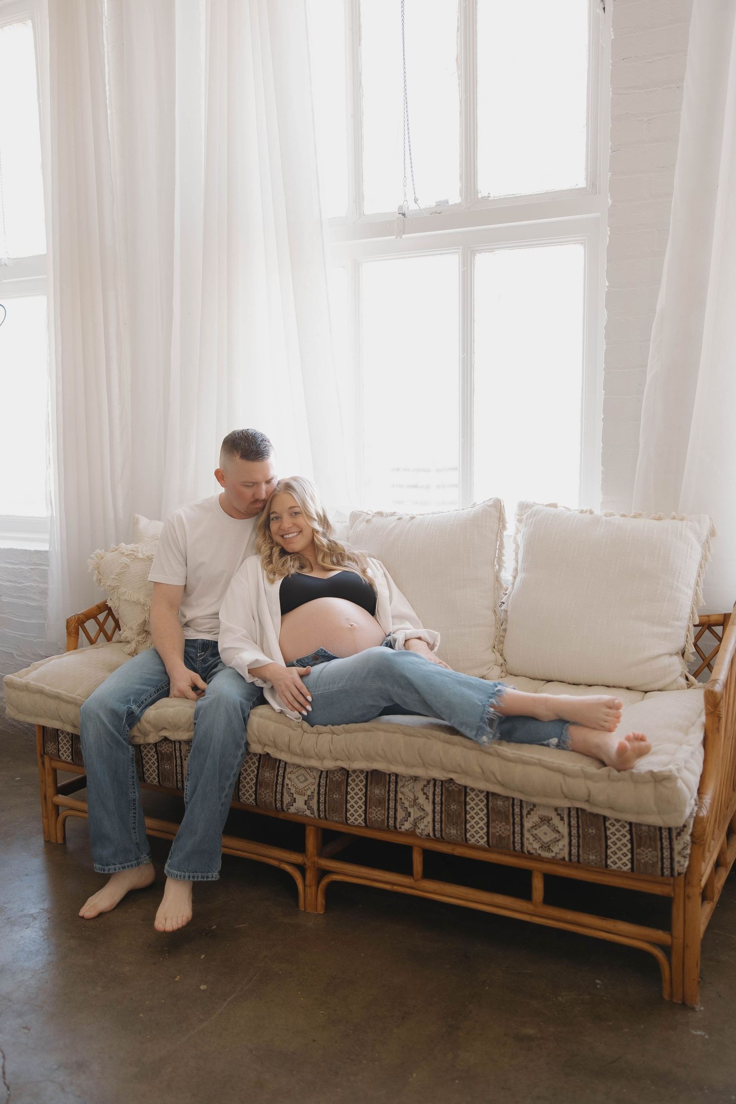 Expectant couple relaxes together on rattan daybed with white cushions near bright window with flowing curtains.