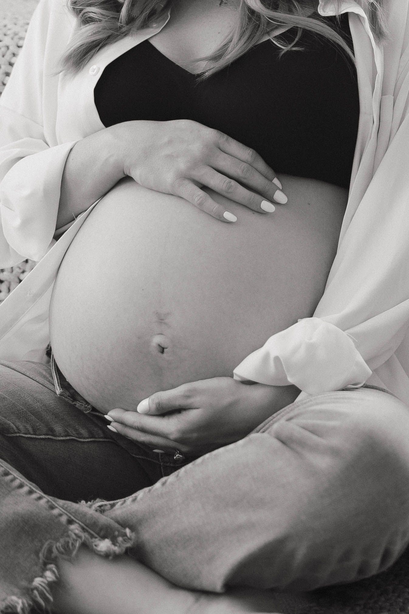 Black and white maternity photo showing pregnant belly with hands gently resting on bare bump in soft natural lighting.