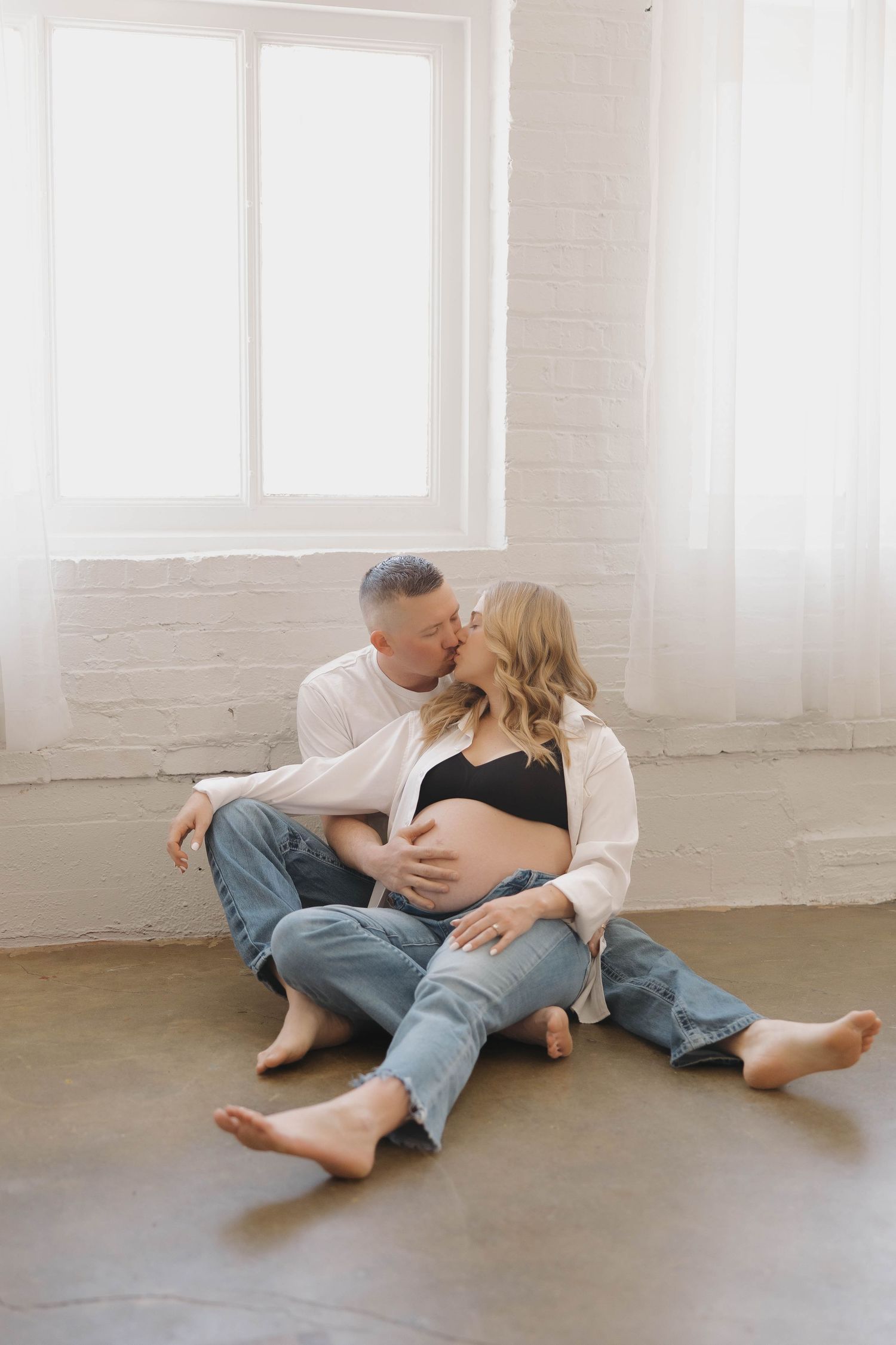 Intimate couple in casual clothing embraces while sitting on concrete floor in bright minimalist studio space.