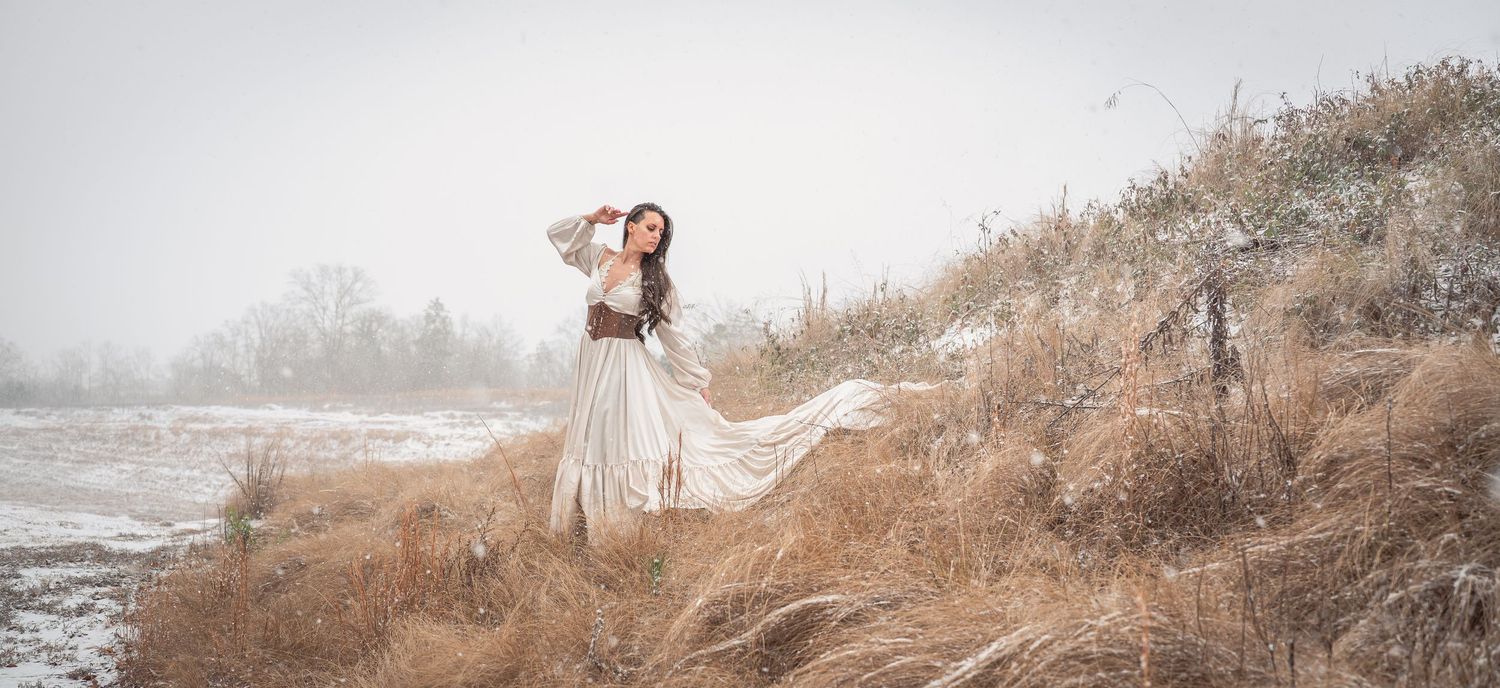 A dreamy winter landscape photo shows a person in a flowing white dress walking through a snowy field at dusk.