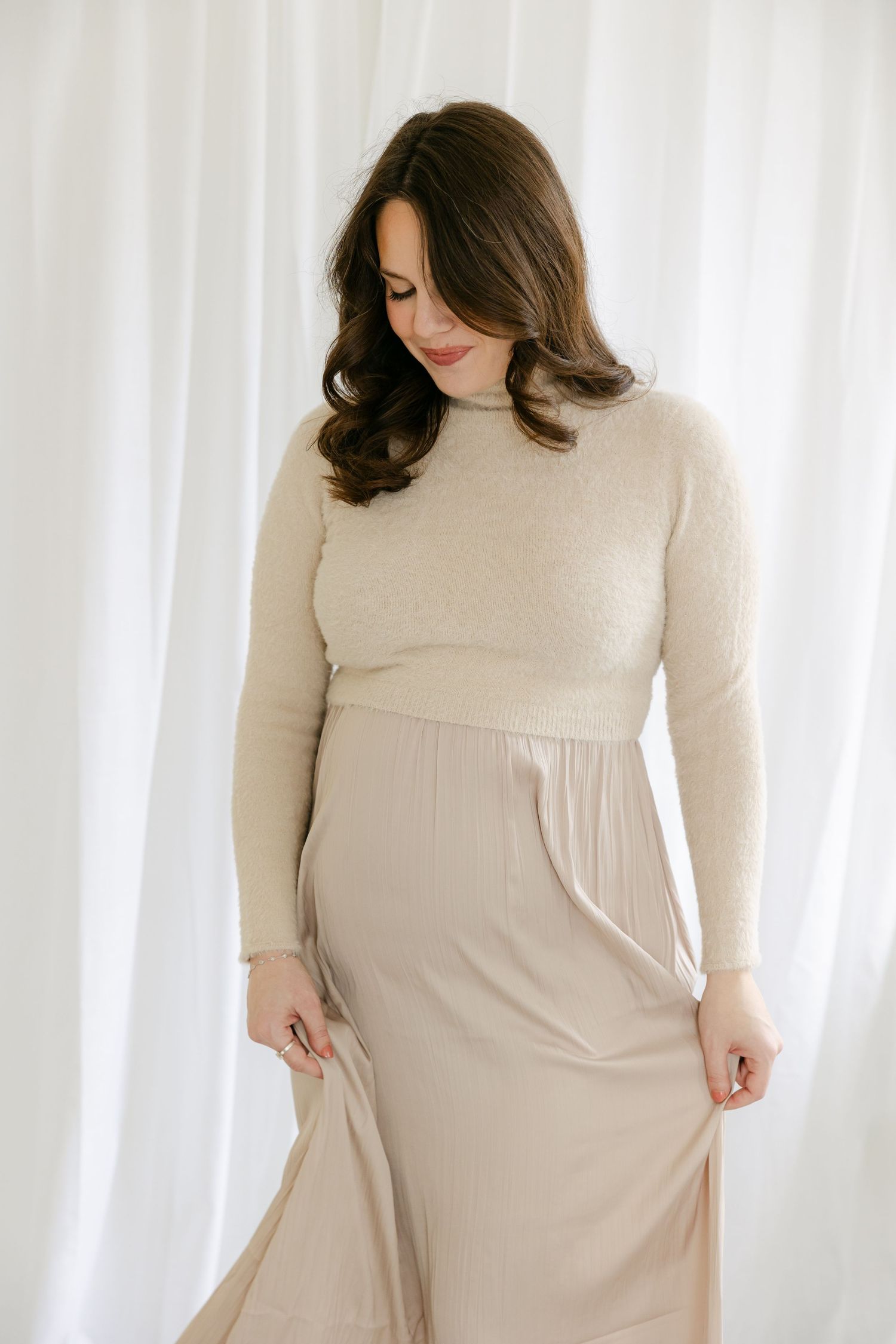 A pregnant woman in a cream-colored sweater dress poses against a white curtain backdrop. Hartford Newborn Photographer, Ashley Nielsen