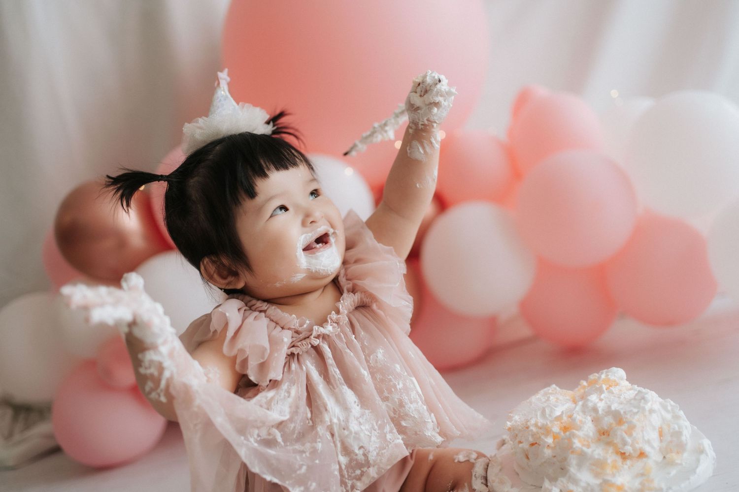 A baby in a white lace dress smiles and plays among pink and white balloons at a festive celebration.