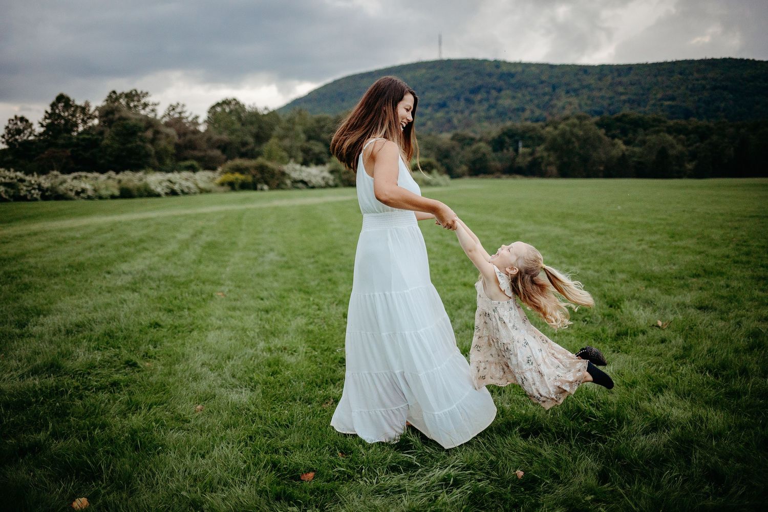 A person in a flowing white dress twirls gracefully in a lush green meadow with mountains in the background.