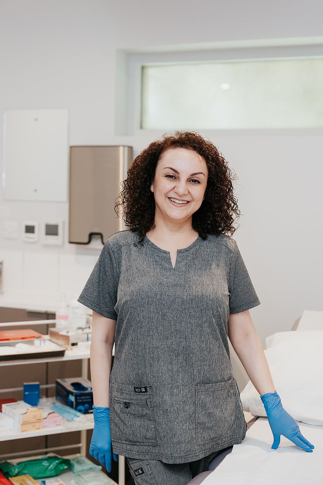 Healthcare worker in gray scrubs and blue gloves stands in a medical facility with a warm expression.