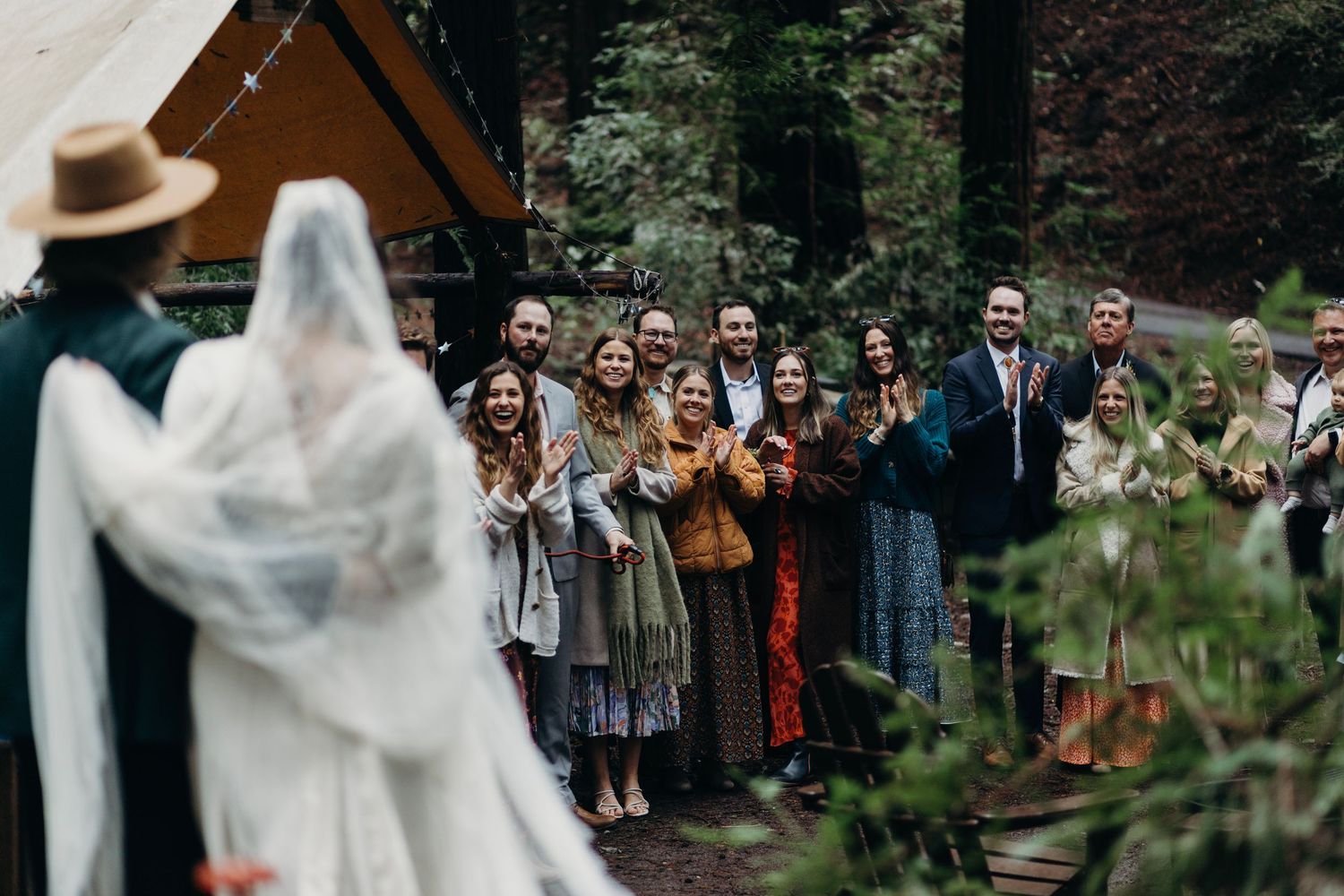 Wedding ceremony guests gather outdoors in a wooded garden setting while watching a couple exchange vows.